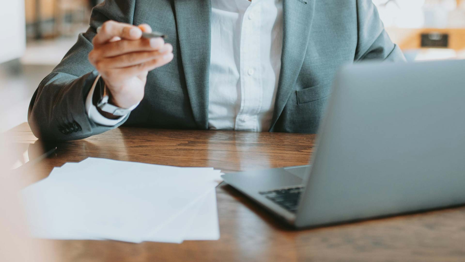 man in suit sitting in front of desk holding up pen in front of laptop and papers